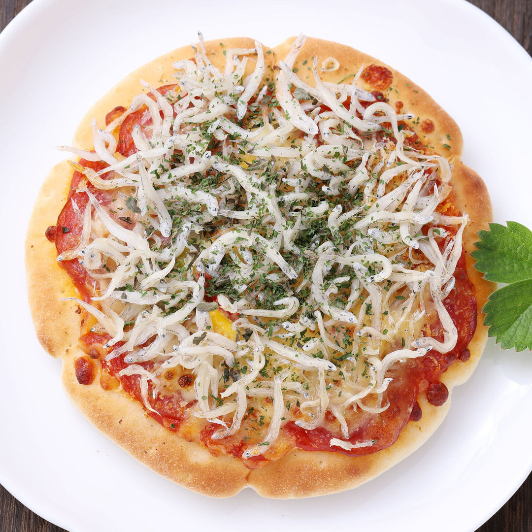 A thin crust pizza topped with Bokksu Tinned Shirasu Anchovies with Black Pepper and Basil-Infused Oil (2-Pack), tomato sauce, and green herbs, finished with a hint of black pepper, served on a white plate.