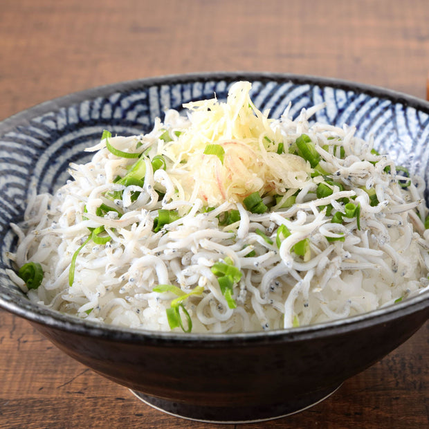 A bowl of white rice topped with Bokksu Tinned Shirasu Anchovies with Black Pepper and Basil-Infused Oil, along with chopped green onions and shredded ginger, sits on a wooden table, delivering a delicate umami flavor in every bite.