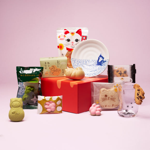 The Cat Lover’s Box showcases a curated assortment of Japanese cat-themed gifts—including snacks, cookies, candies, and adorable ceramics—elegantly displayed on a red box set against a pink background.
