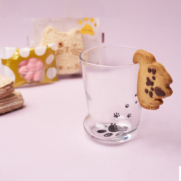 A clear glass with paw prints, holding a cat paw-shaped cookie on its rim, is surrounded by a variety of packaged cat-themed snacks and animal-shaped cookies on a pastel surface—making The Cat Lover’s Box an ideal choice for any Japanese cat gifts collection or for treating a cat enthusiast.