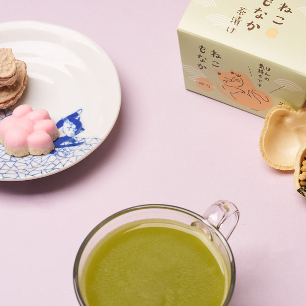 A glass cup of green tea, a plate with pink and beige sweets, and The Cat Lover’s Box – a pastel package featuring Japanese text – all arranged on a light purple surface, perfect for anyone who adores cat-themed snacks and unique Japanese cat gifts.