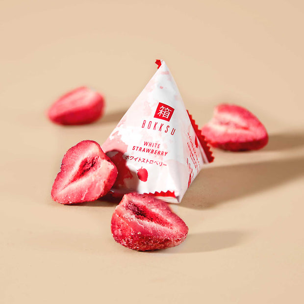 The triangular package labeled "Bokksu Snack Box: Seasons of Japan (FBM)" is surrounded by dried strawberry halves, capturing the essence of Japanese snacks. This delightful treat from Bokksu invites you to savor the flavors of four seasons on a warm beige surface.