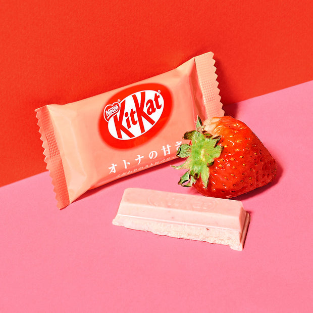 A pink-wrapped KitKat bar labeled in Japanese rests on a pink surface beside a fresh strawberry and an unwrapped pink KitKat piece, showcasing the delicious variety found in the Dagashi Snack Crate—an exciting 30-piece Japanese snack box filled with candy, chocolate, cookies, mochi, and Ramune treats.