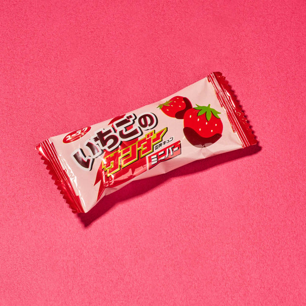 A vibrant pink-wrapped Japanese strawberry chocolate candy bar, adorned with images of strawberries and set against a matching pink backdrop, is just one of the delightful surprises you might discover in the Dagashi Snack Crate—a 30-piece Japanese snack variety box featuring an exciting assortment of candy, chocolate, cookies, mochi, and Ramune treats. Perfect for fans eager to explore unique Japanese snacks and sweets!.