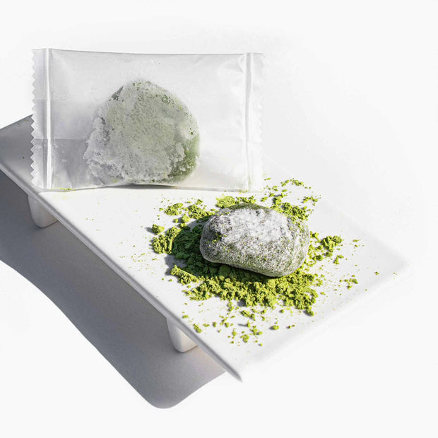 A piece of green mochi from the Dagashi Snack Crate—30-Piece Japanese Snack Variety Box with Candy, Chocolate, Cookies, Mochi & Ramune Treats—partially covered in powdered sugar, sits on matcha powder with another wrapped in clear plastic behind it, arranged on a white rectangular plate.