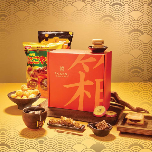 The April '26 Bokksu: Handpicked in Japan snack box is shown featuring a selection of assorted Japanese snacks, sweets, and treats, all neatly arranged on a table with a patterned gold background.